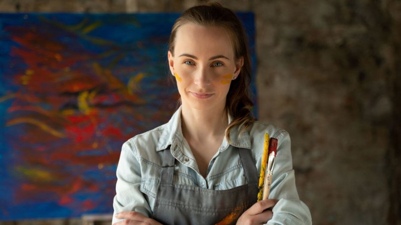 Beautiful Painter Holding Paintbrushes With a Painting at the Back